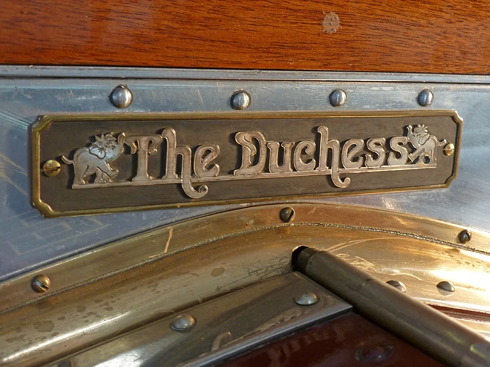 A close-up of the brass nameplate mounted on the firewall of chassis 27NA, engraved 'The Duchess' and flanked by small elephant motifs.