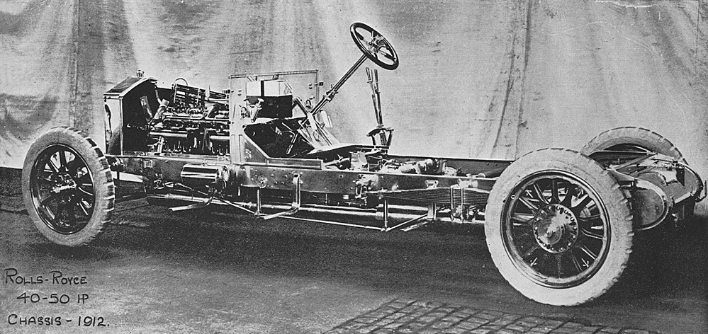 A 1913 Rolls-Royce Silver Ghost rolling chassis — engine, frame, and wooden artillery wheels without any coachwork — photographed against a canvas backdrop for the factory catalogue.